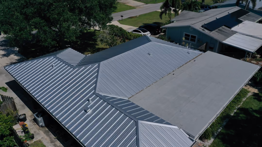 A silver standing seam metal roof seen from the air at an angle, the panel runs and seams unbroken from ridge to eave across the main wing and the rear addition