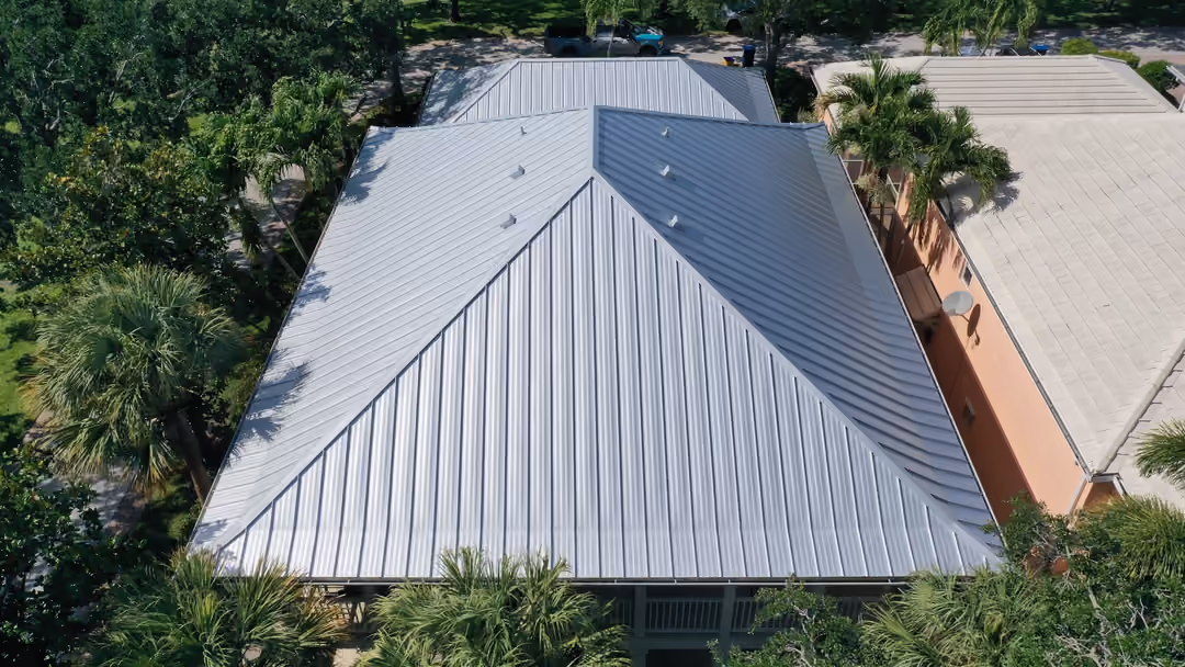 A silver standing seam hip roof seen from above at close range, panel seams running to the ridge, palms and a neighboring tile roof alongside