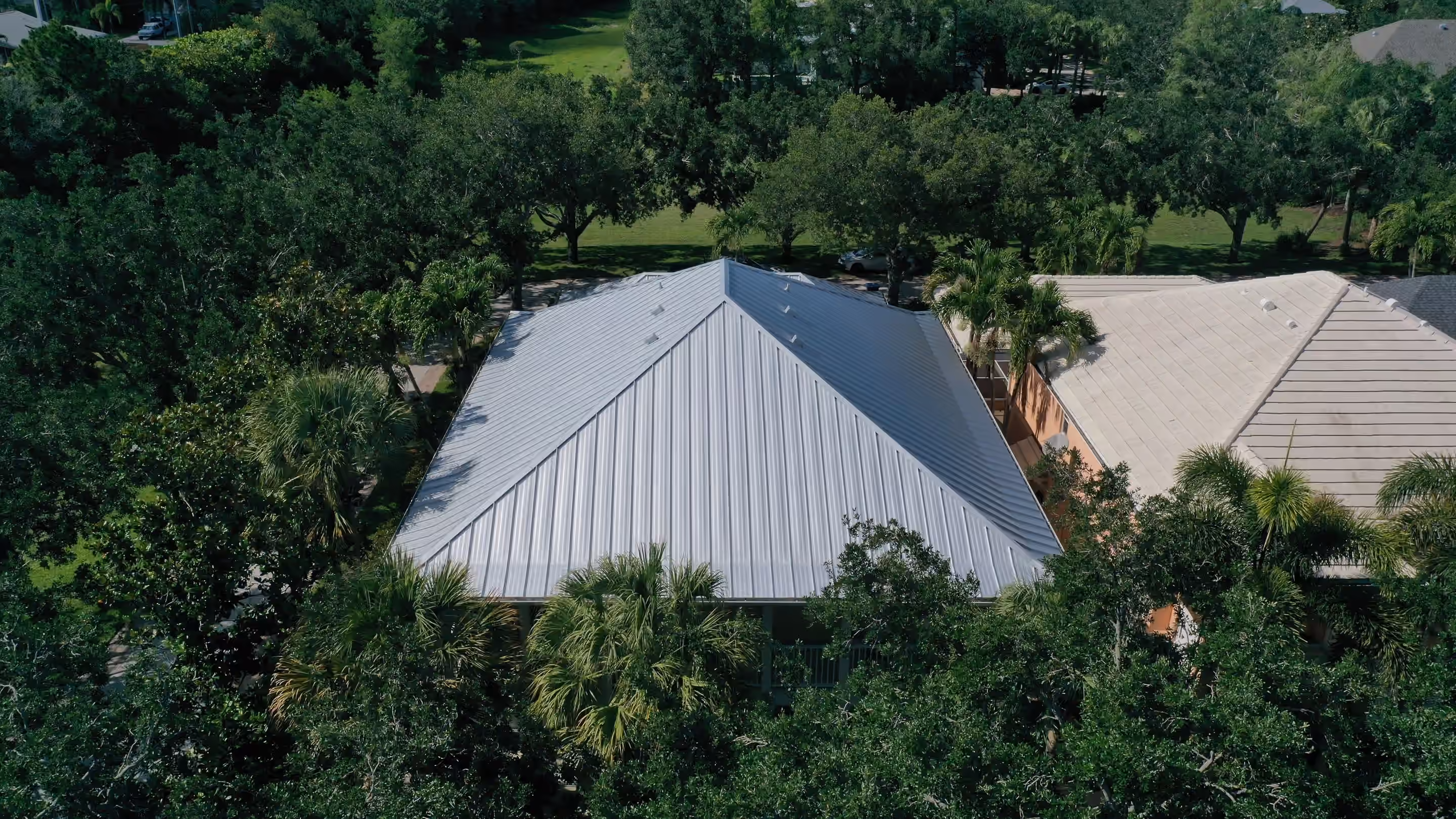 A silver standing seam hip roof set among mature trees, seen from above, with a neighboring tile roof at one side