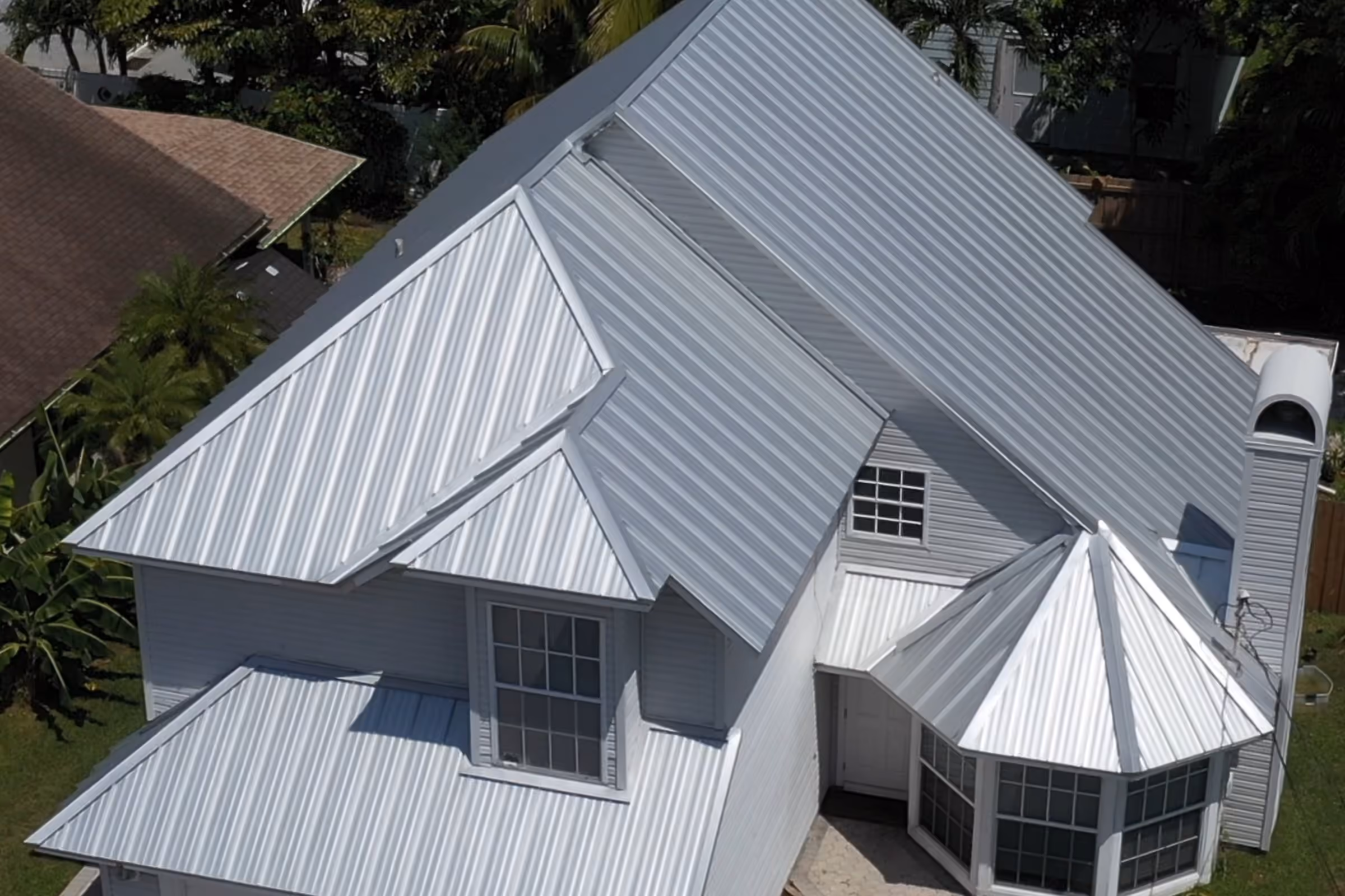 A silver standing seam metal roof over a two-story house seen close from the air, the raised seams running unbroken over the gables and down onto the turret bay at the corner