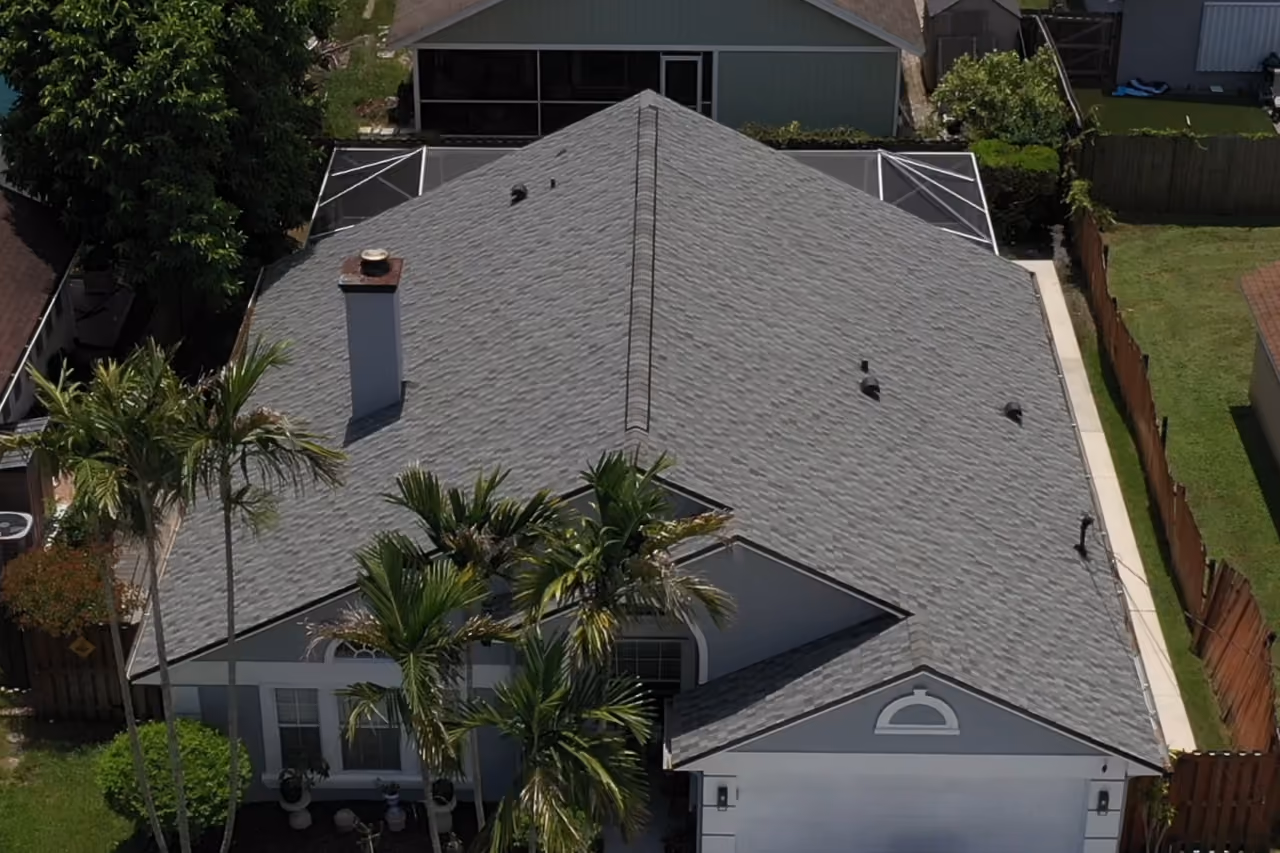 A gray architectural shingle hip roof seen from above, with a chimney on the main slope and a screened pool cage behind the house