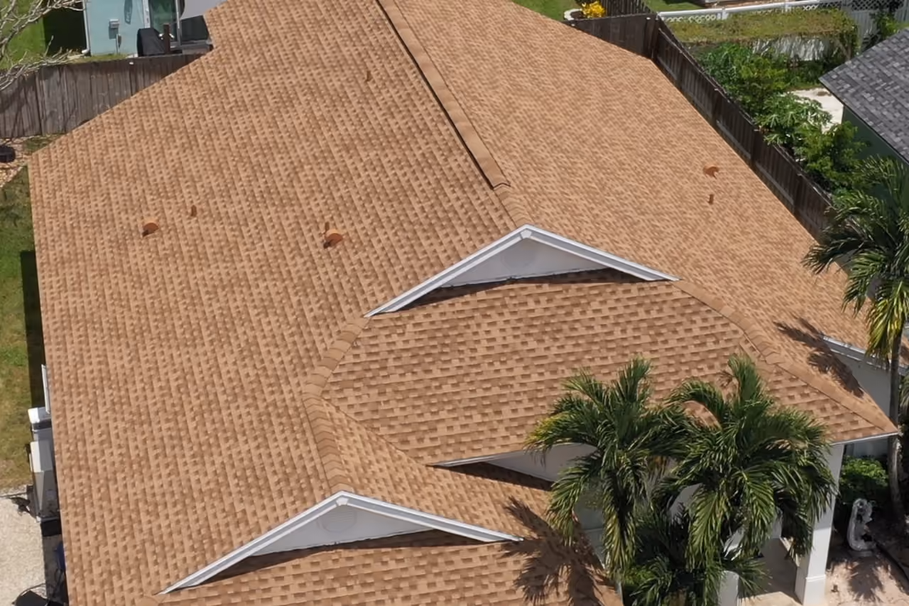A tan architectural shingle hip roof seen from above, with two white gable vents and palms crowding the right edge of the frame