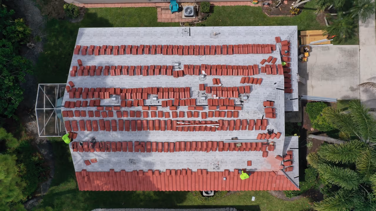 Terracotta tile loaded in rows across the underlayment of a large roof, seen from directly above, with two crew members in high-visibility shirts on the field