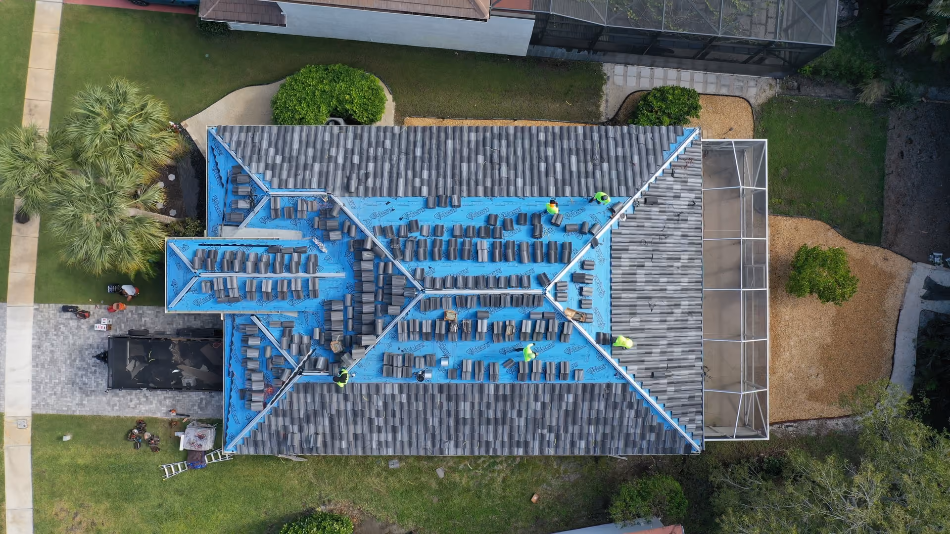 A hip roof mid re-tile seen from directly above, blue underlayment exposed with stacks of gray flat tile staged in rows and finished tile on the far planes