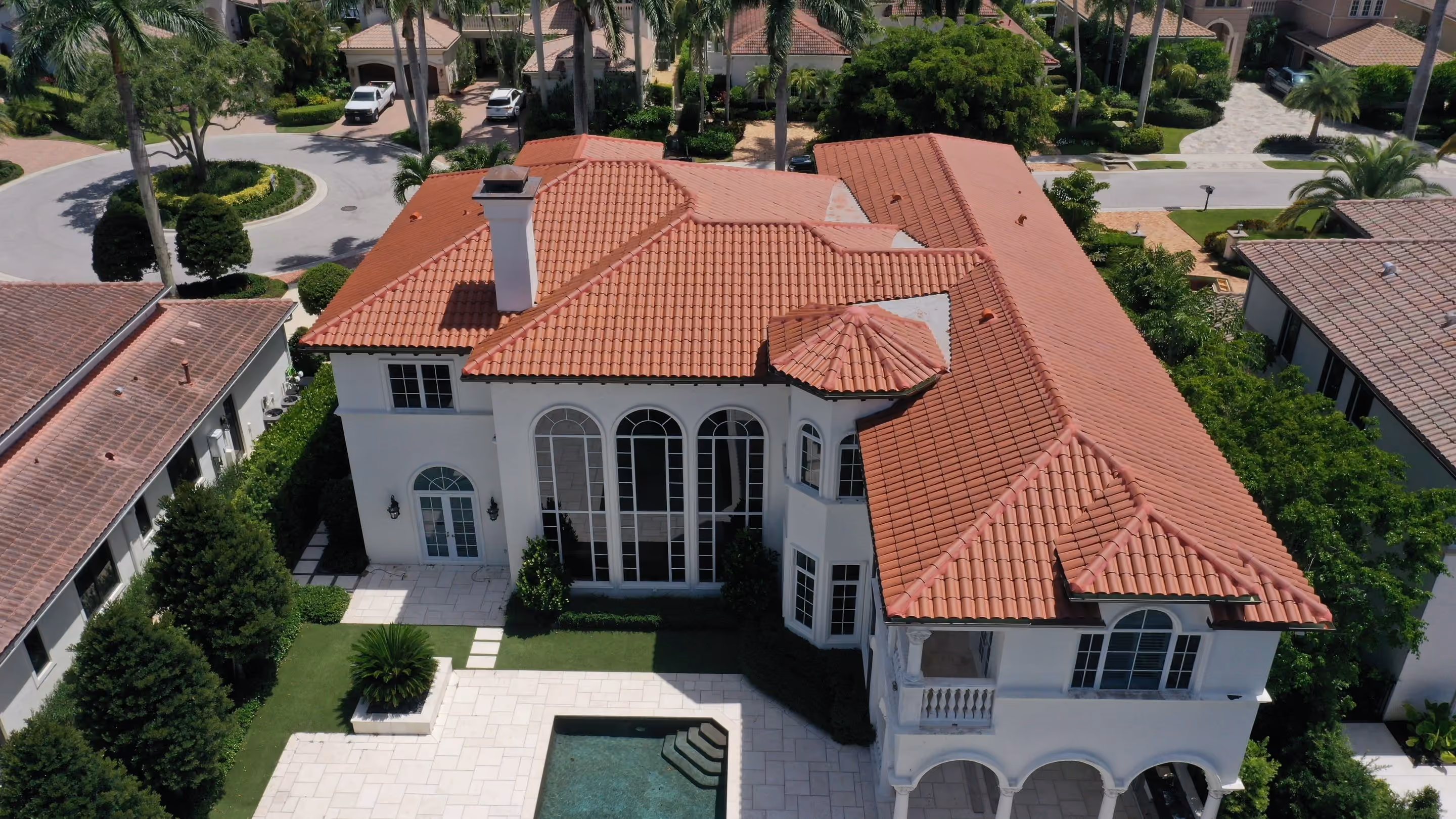 A new terracotta barrel tile roof seen from above on a white Mediterranean estate, its hips and valleys running down past a turret to tall arched windows, with a pool deck behind the house and a palm-planted cul-de-sac in front