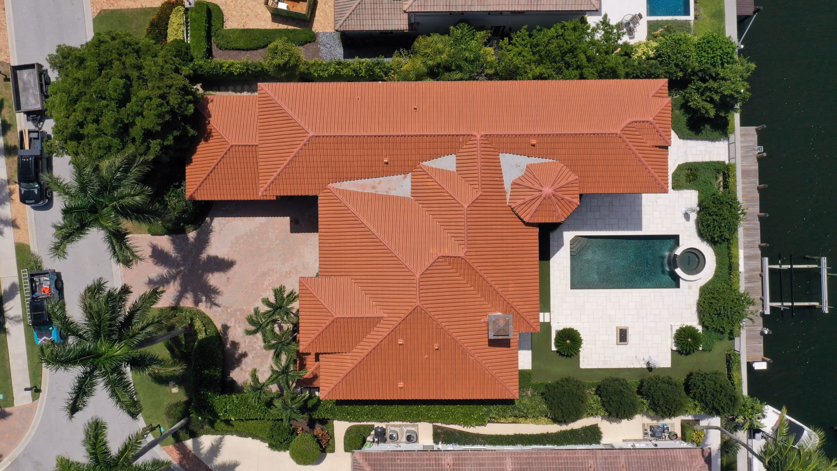 A large terracotta tile roof seen from directly above, its hips meeting around a round turret section, with a pool and a boat dock at the edge of the lot