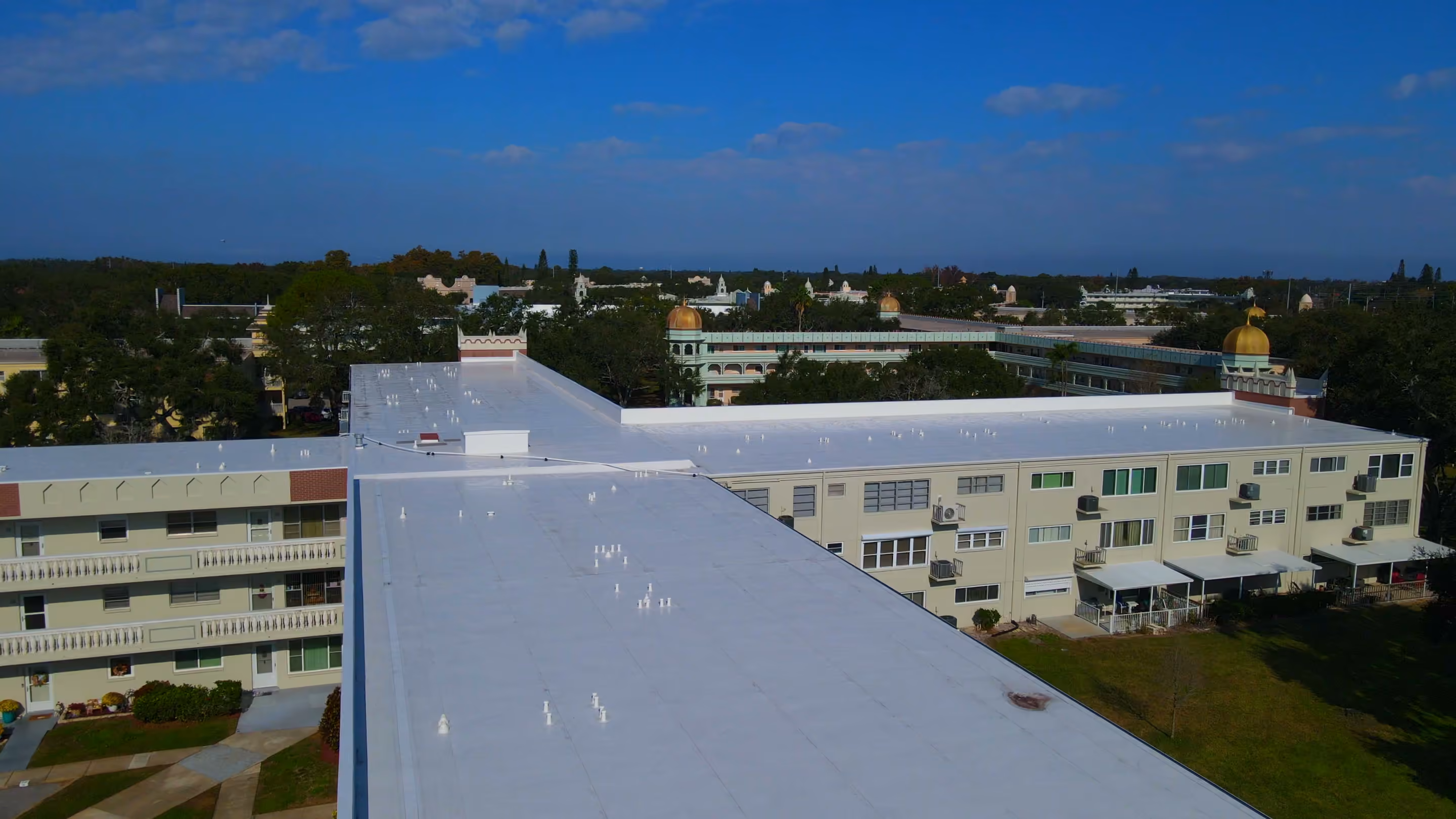 A white flat roof on a three-story apartment building seen from the air, with more flat-roofed buildings and trees behind it under a blue sky.