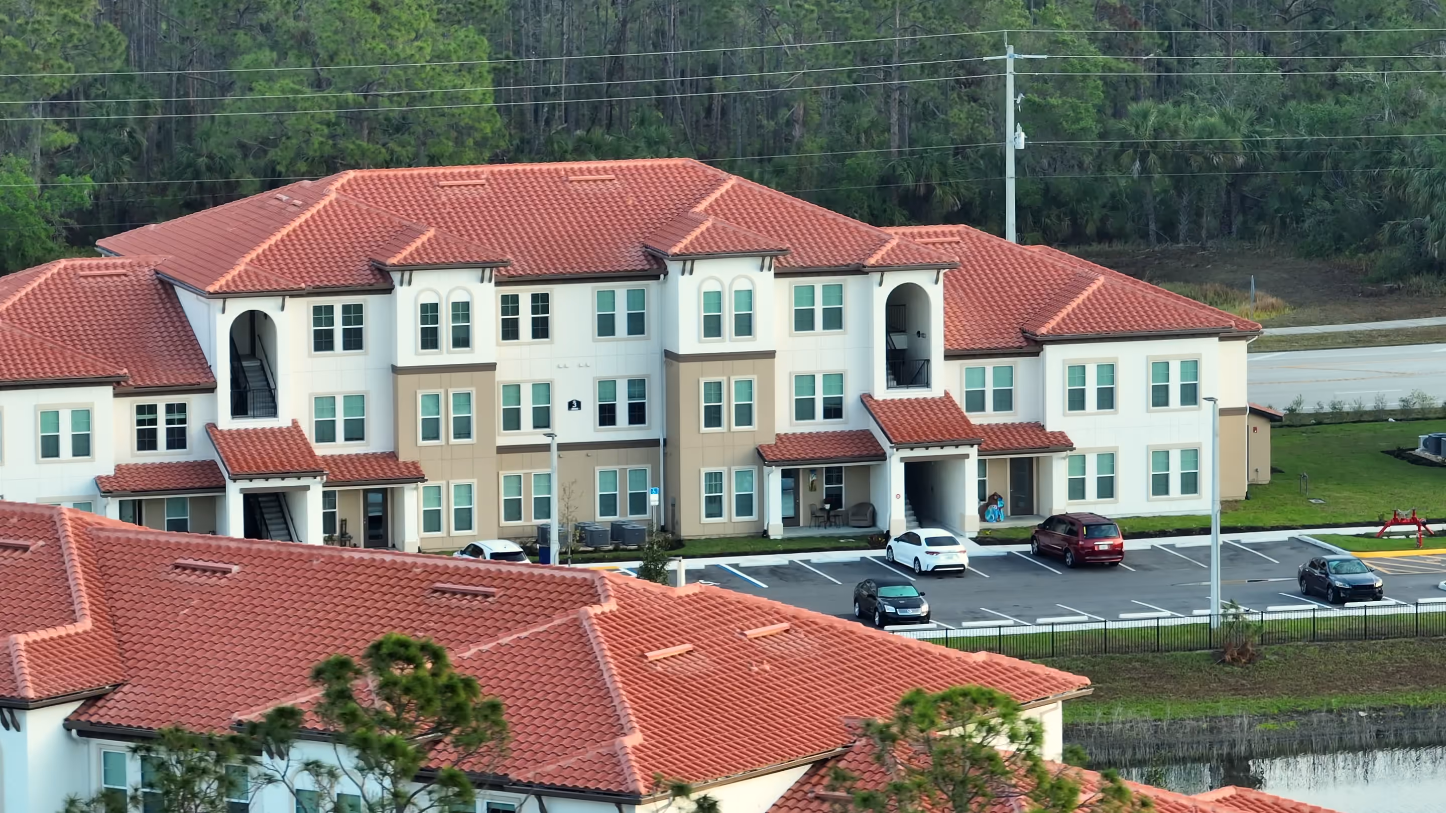 Three-story apartment buildings with red tile roofs seen from the air, with a parking lot beside them and pine woods behind