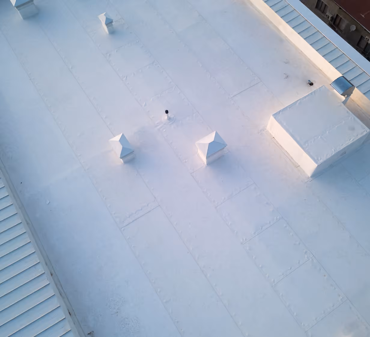 A white flat roof seen from above, with square vent hoods and a raised unit on the roof and standing seam metal along its edges.