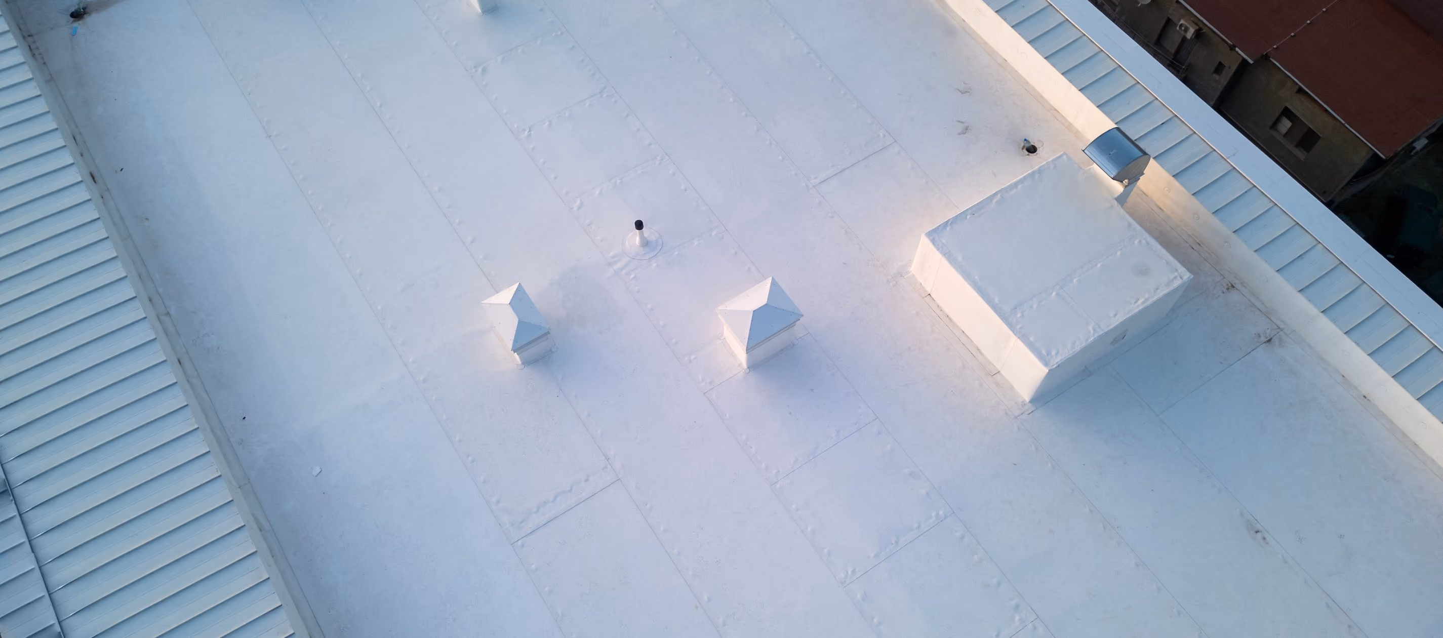 A white flat roof seen from above, with square vent hoods and a raised unit on the roof and standing seam metal along its edges.
