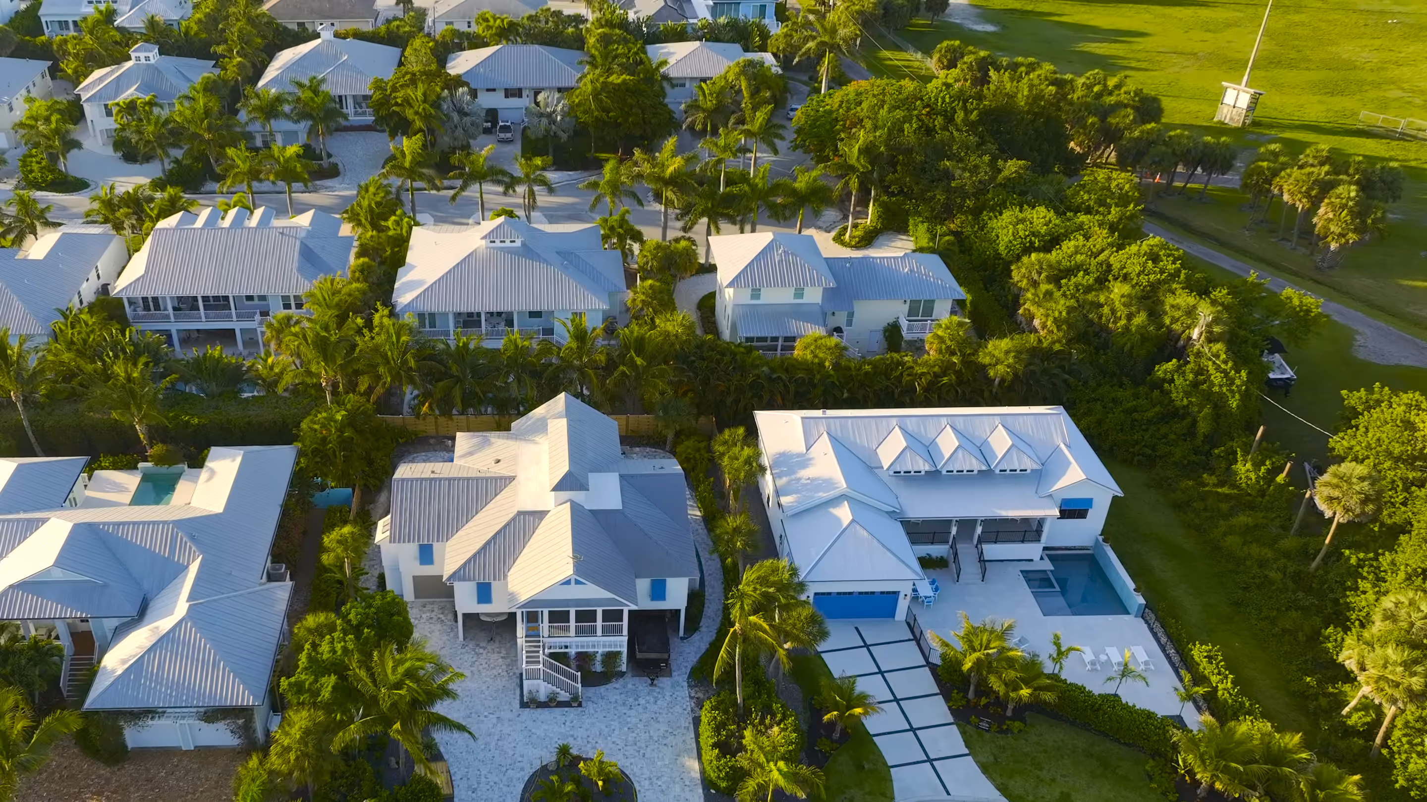 White and gray metal roofs seen from the air, with palm trees between them and a green field beyond the trees